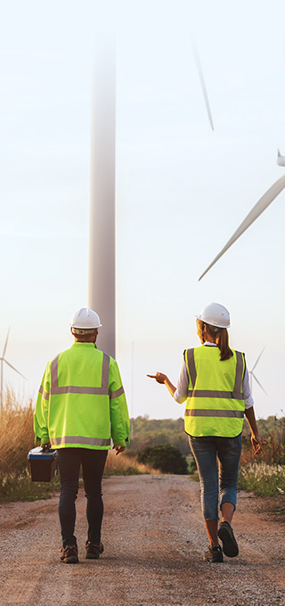 Turbine technician in neon safety jacket standing on the top of the hill and looking at the perspective of hills covered with greenery and wind turbines above them.